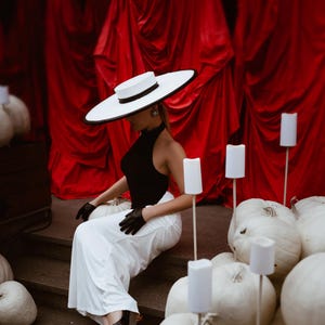 May include: A woman in a black top, white pants, and a wide-brimmed hat sits on steps surrounded by white pumpkins. The background features red curtains and white candle-shaped lights on sticks. She wears black gloves and shoes.