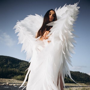 May include: A woman wearing a white feathered angel costume with large wings. She is standing in a rocky riverbed with a green hillside in the background.