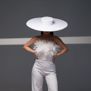 May include: A white wide-brimmed hat and a strapless top adorned with white feathers. The model is wearing a light gray jumpsuit. The outfit is set against a gray backdrop, creating a minimalist fashion statement.