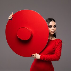 Puede incluir: Una mujer con un vestido rojo sostiene un gran sombrero redondo rojo frente a ella. El sombrero está ligeramente inclinado hacia un lado y la mujer mira hacia un lado.