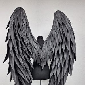 Puede incluir: Grandes alas de ángel de color gris oscuro con un diseño de plumas puntiagudas superpuestas. Las alas están montadas sobre un torso de maniquí negro, mostrando una amplia envergadura con segmentos en forma de pluma dispuestos simétricamente.
