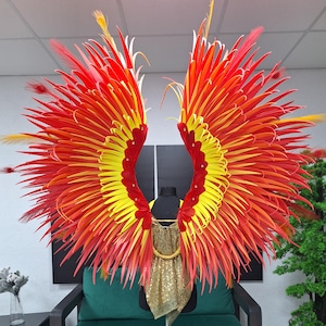 May include: A vibrant costume piece featuring large, feathered wings in shades of red, orange, and yellow. The wings are attached to a gold sequined bodice, creating a dramatic and eye-catching effect. The wings are displayed on a mannequin.