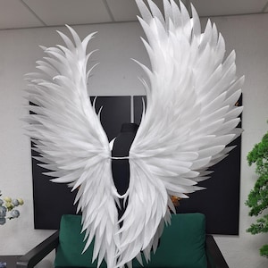 May include: Large, white angel wings with layered, feather-like details. The wings are displayed on a black mannequin, mounted on a silver stand. The wings are the focal point, with a dark green sofa and a black background.
