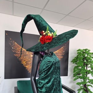 May include: A dark green velvet witch hat with a wide brim, decorated with red roses, berries, and foliage. The hat is part of a costume, worn with a matching green velvet cloak and a purple dress. The outfit is displayed on a black mannequin.
