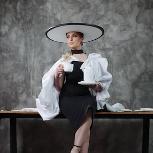 May include: A woman in a black dress and white cape, wearing a large white hat with a black trim, holding a teacup and saucer. She is seated on a dark wooden table. The background is a grey wall.