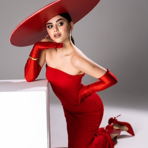 Peut inclure: Une femme vêtue d'une robe rouge sans bretelles, de gants assortis et de talons hauts. Elle porte également un grand chapeau rouge à larges bords et des bijoux dorés. La femme est agenouillée près d'un cube blanc.