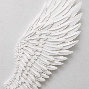 May include: A white, sculpted wing with detailed feather textures. The wing is a decorative piece, possibly for wall art or home decor. The feathers are layered, creating depth and a sense of movement. The overall design is clean and elegant.