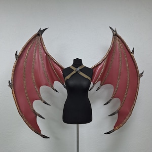 May include: Red and gold dragon wings with a black mannequin. The wings have a red membrane with gold trim and black claw-like tips. The mannequin is wearing a harness. The wings are designed to be worn as a costume.