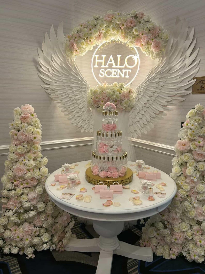 May include: A display for Halo Scent featuring a tiered perfume bottle arrangement on a round white table. The table is decorated with pink handbags, rose petals, and small perfume bottles. The backdrop includes a neon sign with the Halo Scent logo, angel wings, and floral arrangements.