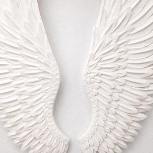 May include: A pair of white angel wings, each with detailed feather textures, are displayed against a plain white background. The wings are symmetrical, with a curved shape and a layered design, creating a sense of depth and dimension.