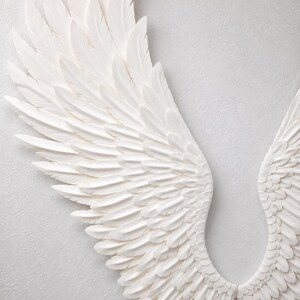 May include: A pair of white, textured angel wings, with detailed feathering. The wings are mounted on a light gray wall, creating a dimensional effect. The feathers have a slight curve, adding to the realistic appearance.