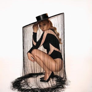 May include: A black bodysuit with a geometric design and a matching black hat. The model is wearing black high heels. The background is white. The outfit is framed by a black fringe.