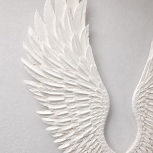 May include: Two white, textured angel wings are displayed against a light gray background. The wings are detailed with individual feather carvings, creating a sense of depth and realism. The wings are a decorative item.