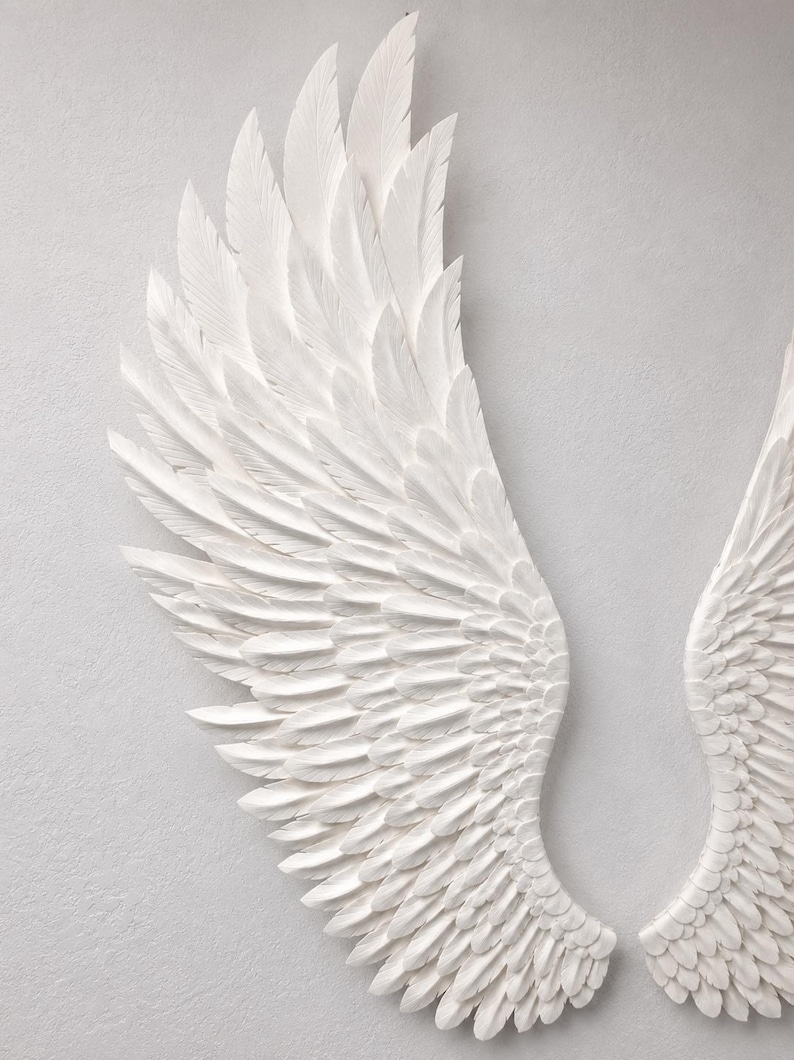 May include: Two white, textured angel wings are displayed against a light gray background. The wings are detailed with individual feather carvings, creating a sense of depth and realism. The wings are a decorative item.
