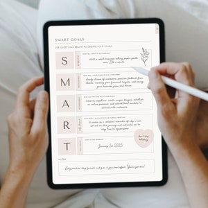以下が含まれることがあります： ピンクの背景に手書きのメモが書かれたSMART目標ワークシートが表示されているデジタルタブレット。ワークシートには、「具体的」「測定可能」「達成可能」「関連性」「時間制限」など、目標設定に役立つ質問のチェックリストが含まれています。ワークシートには、メモ欄と「信じ続ける」というテキストが書かれたハート型のステッカーも含まれています。
