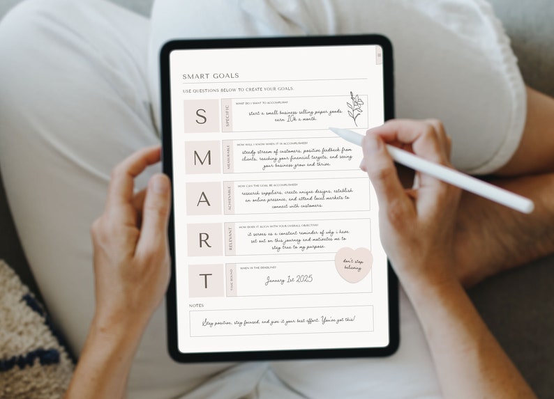 以下が含まれることがあります： ピンクの背景に手書きのメモが書かれたSMART目標ワークシートが表示されているデジタルタブレット。ワークシートには、「具体的」「測定可能」「達成可能」「関連性」「時間制限」など、目標設定に役立つ質問のチェックリストが含まれています。ワークシートには、メモ欄と「信じ続ける」というテキストが書かれたハート型のステッカーも含まれています。