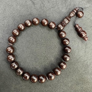 May include: A dark brown prayer bead bracelet with a tassel. The beads are round and polished, with a visible wood grain pattern. The bracelet is laid out on a gray surface.