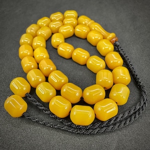May include: A prayer bead strand featuring smooth, barrel-shaped yellow beads arranged in a continuous loop. The beads are strung on a black braided cord that extends into a tassel at one end, with a single bead attached to the end of the cord.