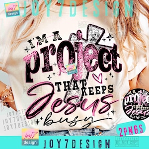 May include: Cream-colored t-shirt with the text "I'M A project THAT KEEPS Jesus busy" in black and pink. The design includes a cross, hearts, and stars. The shirt also has the text "2 PNGS" in the bottom right corner.