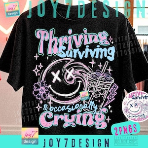 May include: Black t-shirt with a graphic design featuring the words "Thriving Surviving & occasionally Crying" in pink and blue. The design includes a smiley face, a flower, and a drink with ice.