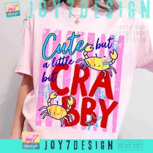 May include: Pink t-shirt with a graphic design featuring the text "Cute but a little bit Crabby" in blue, red, and yellow. The design includes two yellow crab illustrations and blue star accents.