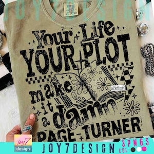 May include: A sage green t-shirt with the text "Your Life YOUR PLOT make it a damn PAGE-TURNER" in distressed black lettering. The design includes an open book with floral illustrations and lightning bolts. The shirt has a crew neck and short sleeves.