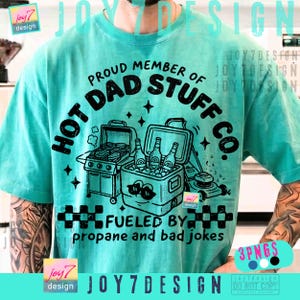 May include: Turquoise t-shirt with a graphic design. The design includes the text "HOT DAD STUFF CO." and "PROUD MEMBER OF." Below the text is a grill, cooler with bottles, and a lawnmower. The text "FUELED BY propane and bad jokes" is also included.