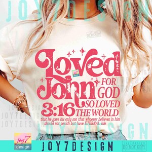 Op de afbeelding: Cr&egrave;mekleurig T-shirt met de tekst "Loved for John 3:16" in een vetgedrukte, roze lettertype. Het ontwerp bevat sterren en de zin "So loved the world" in een gestileerd schrift. Het shirt bevat ook een bijbelvers.