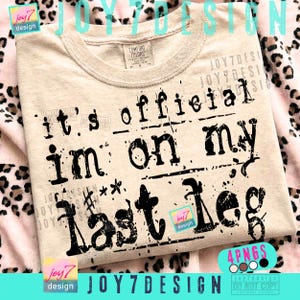 May include: A beige Comfort Colors t-shirt with the phrase "it's official I'm on my last leg" printed in black. The design includes stamp-like text and decorative elements. The shirt has a crew neck.