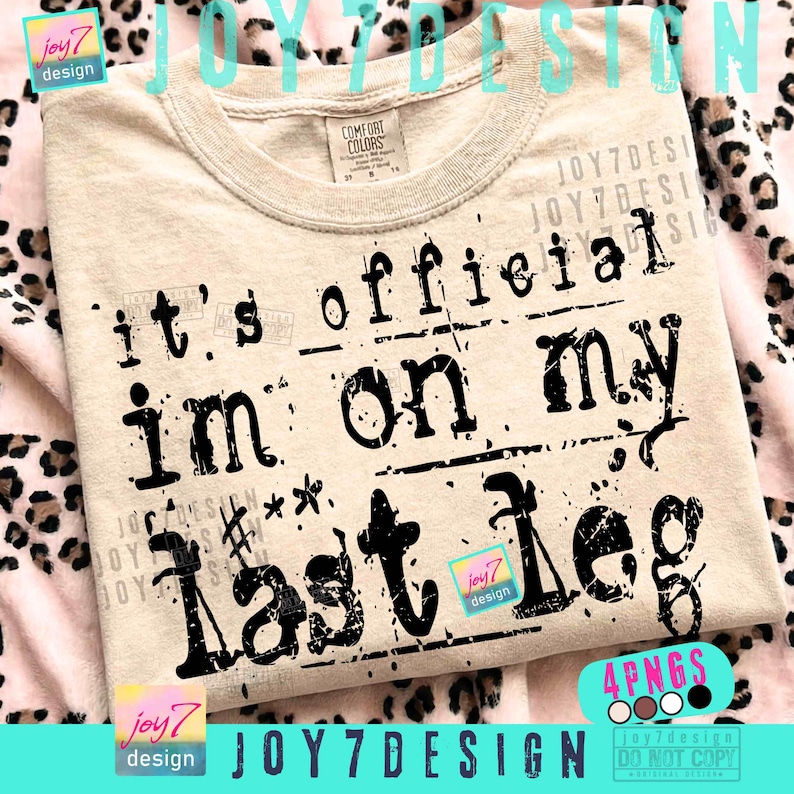 May include: A beige Comfort Colors t-shirt with the phrase "it's official I'm on my last leg" printed in black. The design includes stamp-like text and decorative elements. The shirt has a crew neck.