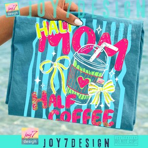 May include: Teal t-shirt with a colorful graphic that reads "Half Mom Half Coffee." The design includes a coffee cup, bows, and stars. The text and images are in bright pink, yellow, and green. The shirt is held by a hand.