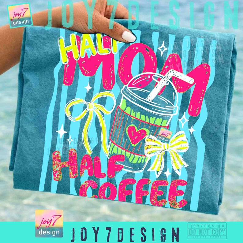 May include: Teal t-shirt with a colorful graphic that reads "Half Mom Half Coffee." The design includes a coffee cup, bows, and stars. The text and images are in bright pink, yellow, and green. The shirt is held by a hand.