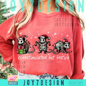 Puede incluir: Sudadera roja con un dise&ntilde;o festivo que presenta tres zarig&uuml;eyas con gorros de Pap&aacute; Noel y luces navide&ntilde;as. El texto "overstimulated but festive" est&aacute; impreso debajo de las zarig&uuml;eyas.