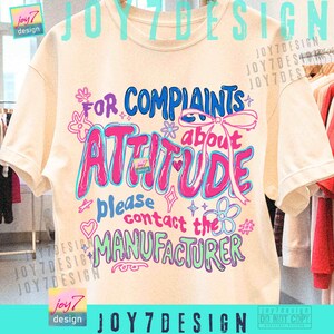 May include: Cream-colored t-shirt with a colorful graphic that reads "For Complaints About Attitude, Please Contact the Manufacturer." The design features pink, blue, and green text with decorative elements. The shirt has short sleeves.