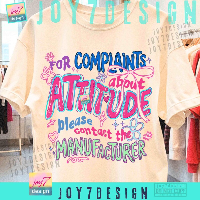 May include: Cream-colored t-shirt with a colorful graphic that reads "For Complaints About Attitude, Please Contact the Manufacturer." The design features pink, blue, and green text with decorative elements. The shirt has short sleeves.