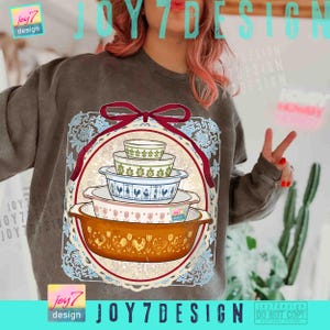 May include: A brown sweatshirt featuring a graphic of a stack of vintage casserole dishes. The dishes are in various colors and patterns, including floral and rooster designs. The graphic is framed by a red ribbon and a decorative border. The text "joy7 design" is visible.
