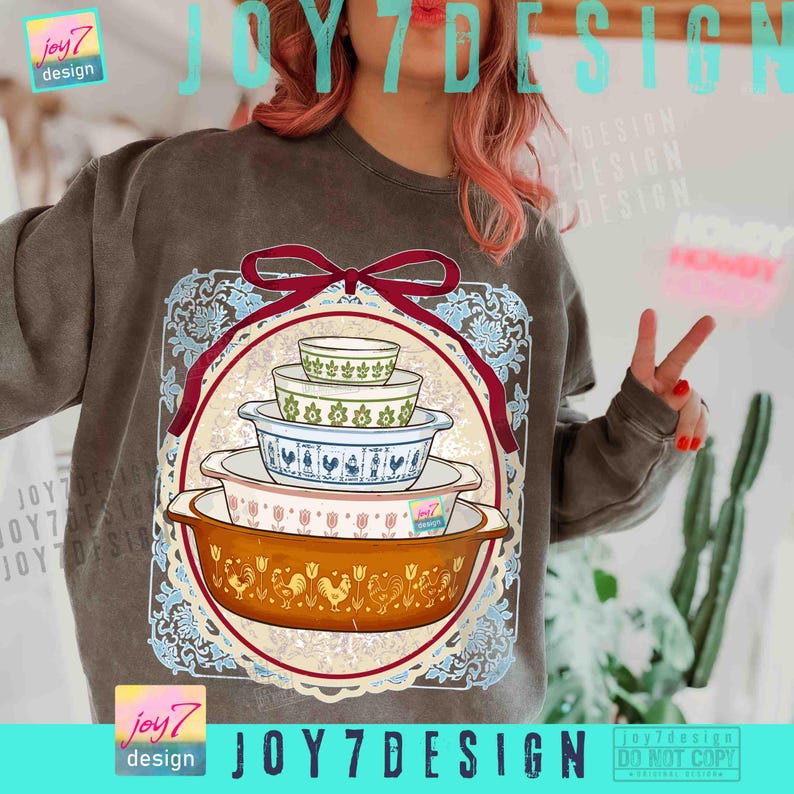 May include: A brown sweatshirt featuring a graphic of a stack of vintage casserole dishes. The dishes are in various colors and patterns, including floral and rooster designs. The graphic is framed by a red ribbon and a decorative border. The text "joy7 design" is visible.