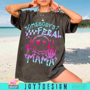 K&ouml;nnte beinhalten: Ein dunkelgraues T-Shirt mit einem grafischen Design, das "Somebody's H*feral Mamas" in Pink, Blau und Wei&szlig; sagt. Die Grafik enth&auml;lt ein Smiley mit gekreuzten Augen und eine Skeletthand, die ein Rock'n'Roll-Zeichen macht.