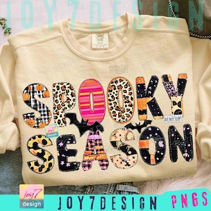 May include: Cream-colored sweatshirt with the words "SPOOKY SEASON" in a patchwork design. The letters feature various patterns, including pumpkins, leopard print, stripes, and bats, perfect for Halloween.