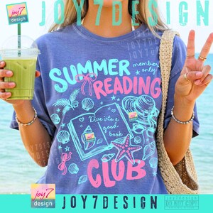 Peut inclure: T-shirt bleu avec un motif graphique rose et turquoise. Le motif comprend les mots "Summer Reading Club", des coquillages, un livre et une &eacute;toile de mer. Le texte "Dive into a good book" est &eacute;galement pr&eacute;sent.