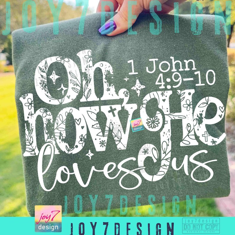 May include: A dark green t-shirt with the words "Oh how He loves us" in white, floral-decorated letters. The text "1 John 4:9-10" is also printed on the shirt. The shirt is held up by a hand with painted nails.