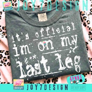 May include: A dark gray Comfort Colors t-shirt with the text "it's official I'm on my last leg" in distressed white lettering. The shirt has a crew neck and short sleeves. The design includes the "joy7 design" logo.