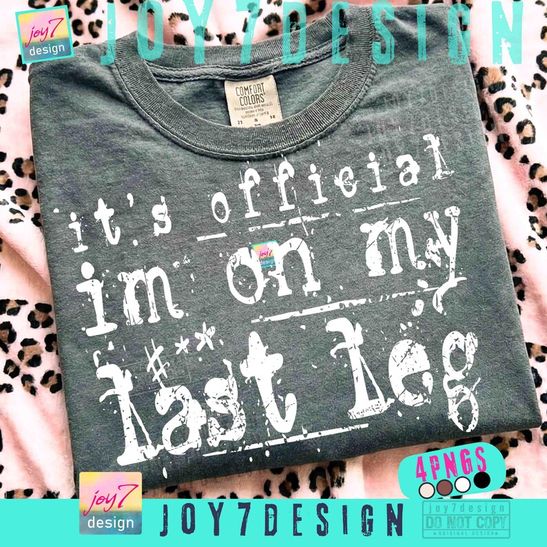 May include: A dark gray Comfort Colors t-shirt with the text "it's official I'm on my last leg" in distressed white lettering. The shirt has a crew neck and short sleeves. The design includes the "joy7 design" logo.
