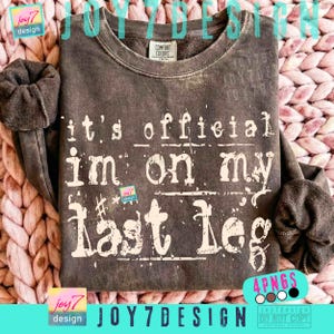 May include: A dark gray sweatshirt with the text "it's official I'm on my last leg" in white lettering. The sweatshirt has a crew neck and long sleeves. The brand name "JOY7DESIGN" is visible in the image.