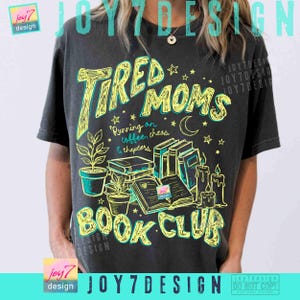 May include: Dark grey t-shirt with a graphic design. The design features the words "Tired Moms Book Club" in yellow and teal, along with illustrations of books, plants, candles, stars, and a crescent moon. The text "Running on coffee, chaos & chapters" is also included.