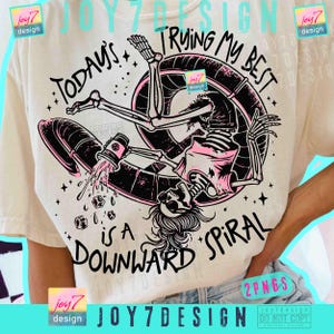 May include: Cream t-shirt with a skeleton graphic inside a pink and black inner tube, holding a mug with liquid spilling out. The text reads "Today's Trying My Best is a Downward Spiral".