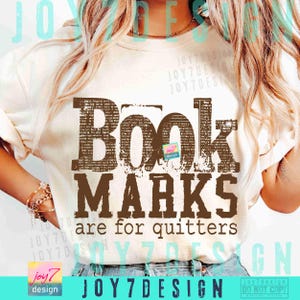 May include: Cream-colored t-shirt with the text "Book Marks are for quitters" in brown, distressed lettering. The shirt features a small logo and the brand name "JOY7DESIGN" at the bottom. The shirt is a casual wear item.