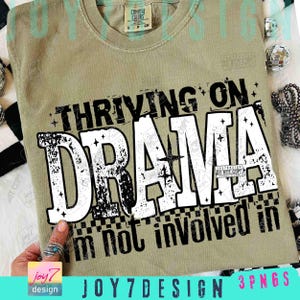 May include: A sage green t-shirt with the text "THRIVING ON DRAMA" in a distressed white font. Below, it reads "I'm not involved in". The shirt has a Comfort Colors tag and a small, square design in the corner.
