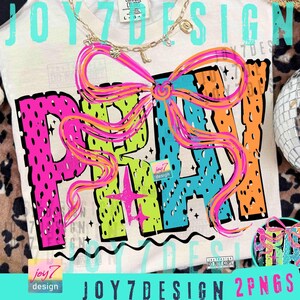 May include: Cream-colored t-shirt with the word "PRAY" in large, colorful letters. The letters are outlined in black and filled with neon green, blue, and orange. A large, pink and orange bow is above the word. A gold necklace is at the top.