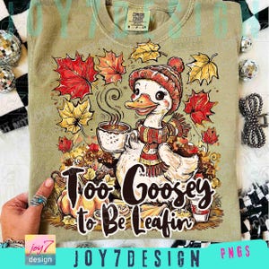 Op de afbeelding: Olijfgroen T-shirt met een herfstthema. Het ontwerp toont een cartoon gans met een hoed en sjaal, die een kopje koffie vasthoudt, omringd door herfstbladeren en pompoenen. De tekst luidt "Too Goosey to Be Leafin".